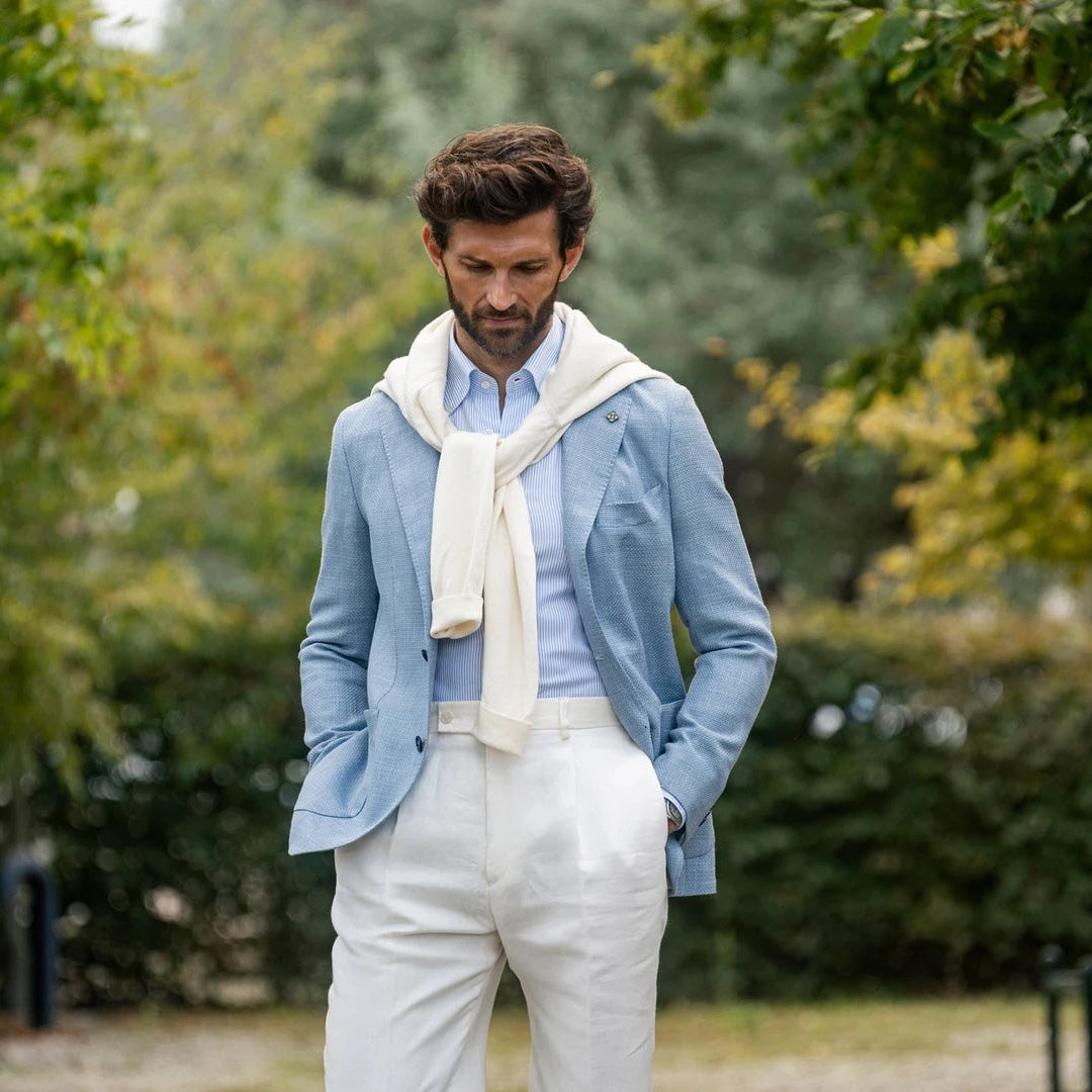 Man wearing a light blue jacket, white shirt, and white pants standing outdoors with greenery in the background