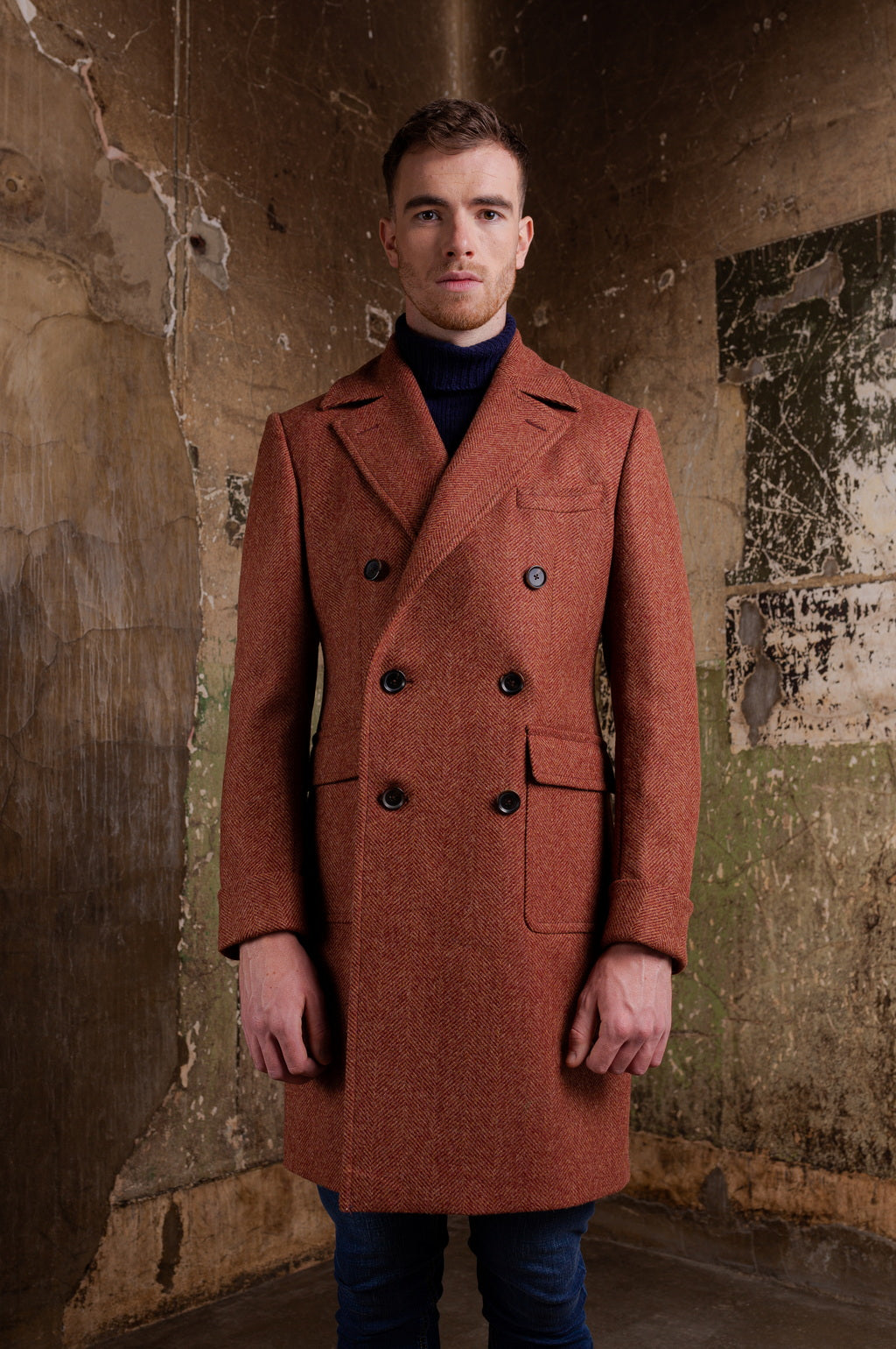 Man wearing a brown coat standing against a textured wall.